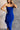 Close up view of model wearing the Liliana Blue Asymmetrical Sleeveless Midi Dress which features royal blue knit fabric, an asymetrical ruffle hem, midi length, a square neckline, and adjustable straps.