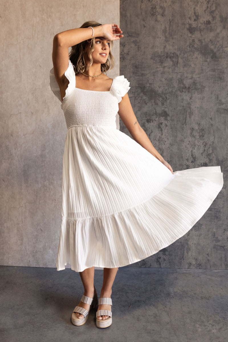 Full body view of model wearing the Katherine Off White Sleeveless Midi Dress which features white fabric with stitched stripe details, a smocked upper with a straight neckline, layered ruffled straps, white lining, and a midi-length hem.
