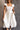 Front view of model wearing the Katherine Off White Sleeveless Midi Dress which features white fabric with stitched stripe details, a smocked upper with a straight neckline, layered ruffled straps, white lining, and a midi-length hem.
