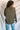 Back view of model wearing the Lost Without You Sweater, which features olive open-knit fabric with larger knitting on the sides, a round neckline, long sleeves, and a slouchy fit.