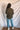 Full body back view of model wearing the Lost Without You Sweater, which features olive open-knit fabric with larger knitting on the sides, a round neckline, long sleeves, and a slouchy fit.