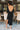 Full body back view of model wearing the Date Night Midi Dress, that has black ribbed knit fabric, a plunge back with pearl and rhinestone trim, a high round neckline, long fitted sleeves, a side slit.