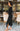 Full body side view of model wearing the Date Night Midi Dress, that has black ribbed knit fabric, a plunge back with pearl and rhinestone trim, a high round neckline, long fitted sleeves, a side slit.