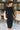 Full body front view of model wearing the Date Night Midi Dress, that has black ribbed knit fabric, a plunge back with pearl and rhinestone trim, a high round neckline, long fitted sleeves, a side slit.