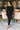 Full body front view of model wearing the Date Night Midi Dress, that has black ribbed knit fabric, a plunge back with pearl and rhinestone trim, a high round neckline, long fitted sleeves, a side slit.
