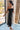 Side view of model wearing the Magic Moment Dress in Black, which features black satin fabric with a midi-length hem, black feathers across the neckline, adjustable thin straps, and a back zipper