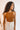 Front view of model wearing the Risk It Bodysuit in Camel, that has stretchy double-layered fabric, a high round neckline, thick straps, and a thong bottom with snap closures. Worn under cream pants