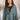 Front view of model wearing the Upstate Twist-Back Top in Green, which has textured green fabric, a button-up front with a collar neckline, long sleeves, and a twist in the back with an open slit. Worn half buttoned.