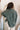 Back view of model wearing the Upstate Twist-Back Top in Green, which features textured green fabric, a button-up front with a collar neckline, long sleeves, and a twist in the back with an open slit