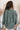 Back view of model wearing the Upstate Twist-Back Top in Green, which features textured green fabric, a button-up front with a collar neckline, long sleeves, and a twist in the back with an open slit