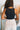 Back view of model wearing the Risk It Bodysuit in Black, that has stretchy double-layered fabric, a high round neckline, thick straps, and a thong bottom with snap closures. Worn under cream pants.