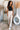 Full body front view of model wearing the Risk It Bodysuit in Black, that has stretchy double-layered fabric, a high round neckline, thick straps, and a thong bottom with snap closures. Worn under cream pants.