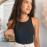 Front view of model wearing the Risk It Bodysuit in Black, that has stretchy double-layered fabric, a high round neckline, thick straps, and a thong bottom with snap closures. Worn under cream pants.