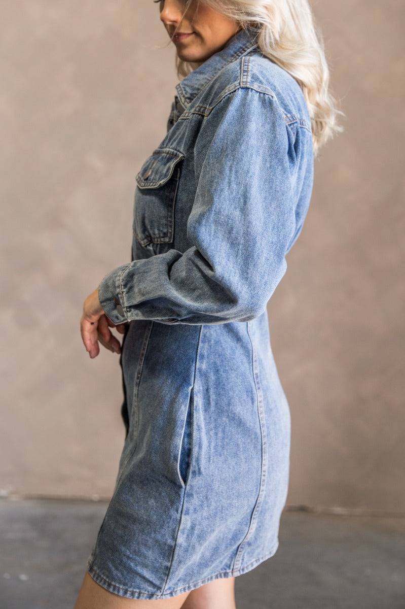 Side view of model wearing the Tessa Blue Denim Long Sleeve Mini Dress which features blue denim fabric, front button up, collared neckline, two front chest buttoned pockets, brown stitching details and long sleeves with buttoned cuffs.