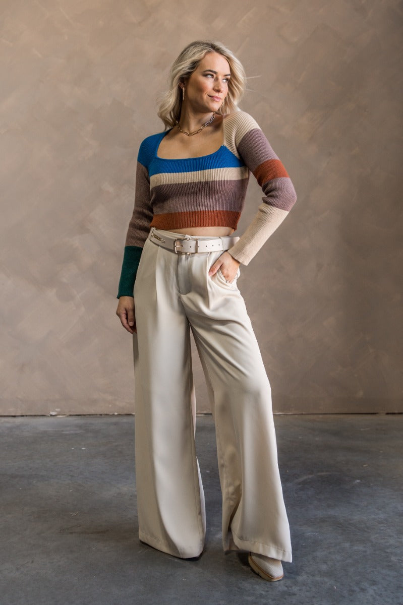 Full body view of model wearing the Natalie Blue Multi Cropped Long Sleeve Top which features taupe, rust, beige and blue knit fabric, striped pattern, cropped waist, square neckline and long sleeves.