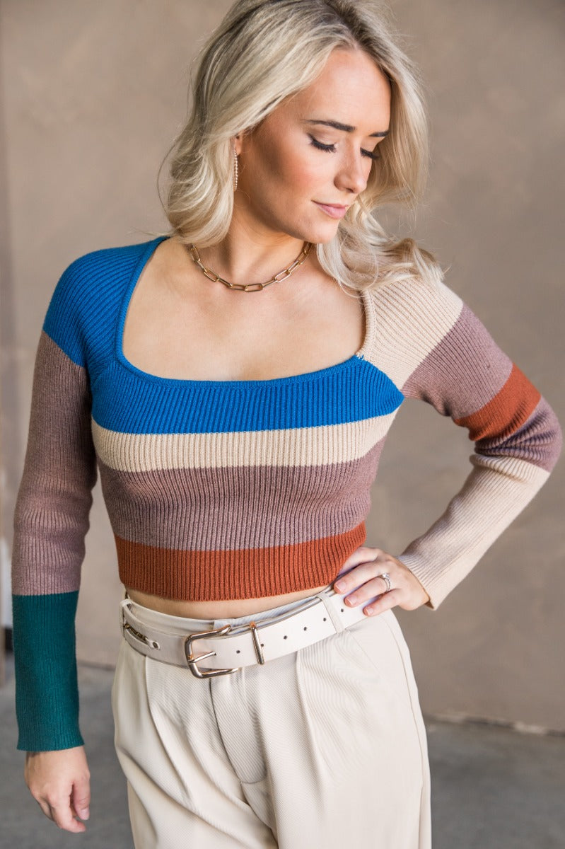 Front view of model wearing the Natalie Blue Multi Cropped Long Sleeve Top which features taupe, rust, beige and blue knit fabric, striped pattern, cropped waist, square neckline and long sleeves.