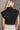 Back view of model wearing the Lola Black Sleeveless Turtleneck Sweater which features black cable knit fabric, a thick hem, a cropped waist, a turtleneck neckline, and a sleeveless design.