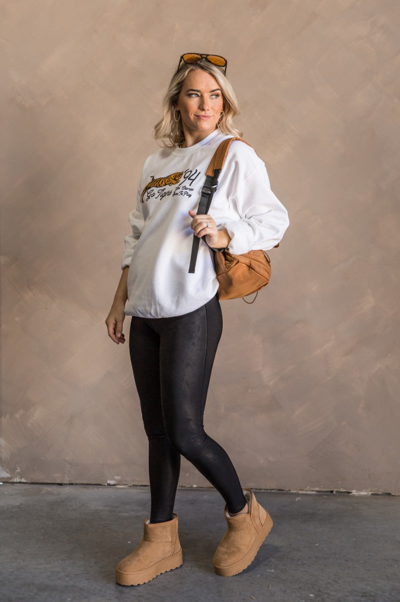 Full body view of model wearing the Girl Champs Tiger Long Sleeve Sweatshirt which features white knit fabric, thick hem, round neckline, black and gold stitch tiger that says "Go Tigers '94, Girl Champs Born To Play" and long sleeves with cuffs.