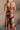 Full body back view of model wearing the Elise Multi Floral Satin Midi Dress which features black, green, pink, orange, turquoise, beige and dark yellow satin fabric, floral print, midi length, cowl neckline, adjustable straps, corset back ties and sleeve