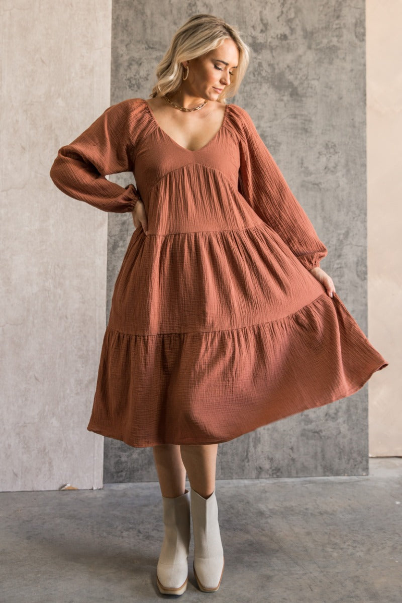 Full body front view of model wearing the Wrenley Rust Long Sleeve Midi Dress that has rust gauze fabric, a tiered body, long balloon sleeves with elastic, side pockets, a v neck, and a smocked back.