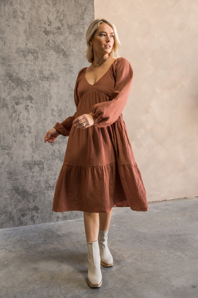 Full body front view of model wearing the Wrenley Rust Long Sleeve Midi Dress that has rust gauze fabric, a tiered body, long balloon sleeves with elastic, side pockets, a v neck, and a smocked back.
