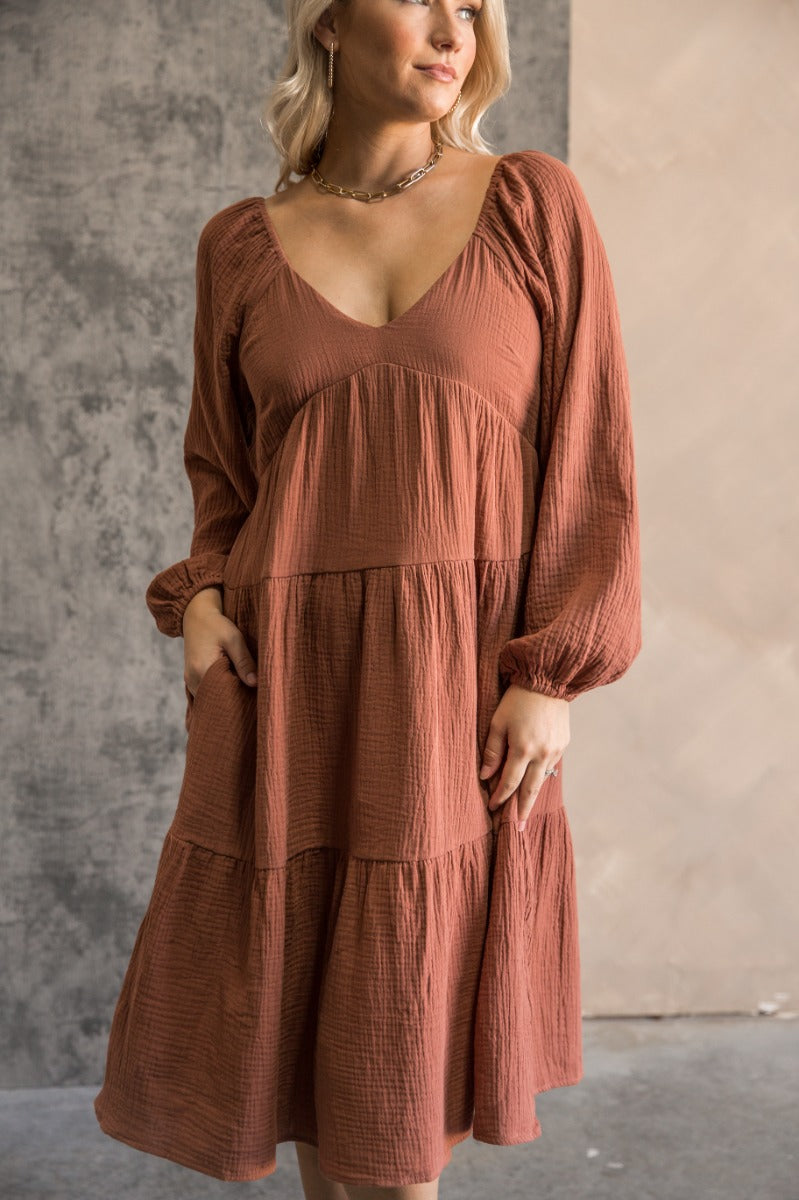 front view of model wearing the Wrenley Rust Long Sleeve Midi Dress that has rust gauze fabric, a tiered body, long balloon sleeves with elastic, side pockets, a v neck, and a smocked back.