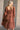 Back view of model wearing the Wrenley Rust Long Sleeve Midi Dress that has rust gauze fabric, a tiered body, long balloon sleeves with elastic, side pockets, a v neck, and a smocked back.