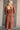 Back view of model wearing the Wrenley Rust Long Sleeve Midi Dress that has rust gauze fabric, a tiered body, long balloon sleeves with elastic, side pockets, a v neck, and a smocked back.