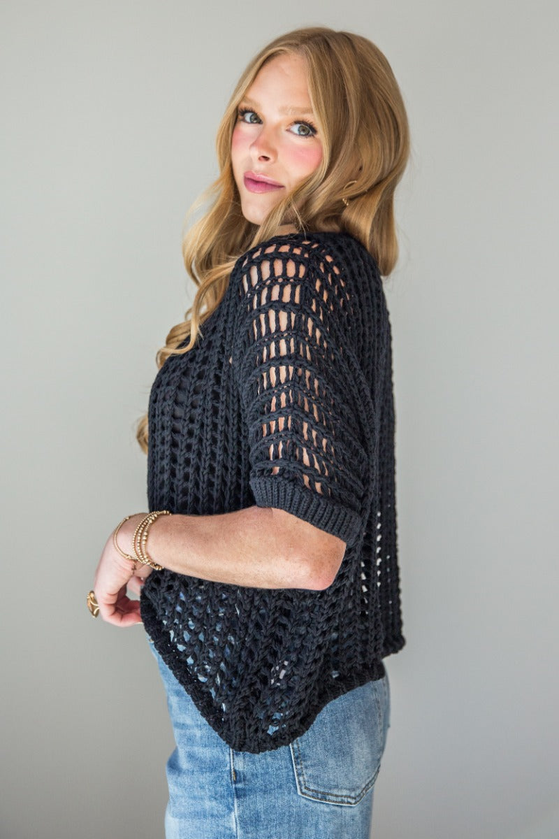 Side view of model wearing the Tatum Black Open Knit Short Sleeve Top which features black crochet knit fabric, round neckline and short sleeves.