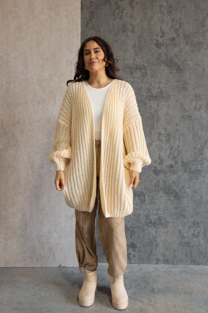 Full body front view of model wearing the Jayla Cream Open Front Cardigan features cream cable knit fabric, an open front, dropped shoulders, a thigh length hem, and long bubble sleeves.