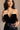 front view of model wearing the Lilliana Black Velvet Strapless Bodysuit that has black velvet fabric, a v-wire strapless neck with ruched details, and a thong bottom. Worn with pants.