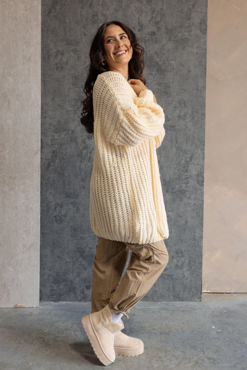 Full body side view of model wearing the Jayla Cream Open Front Cardigan features cream cable knit fabric, an open front, dropped shoulders, a thigh length hem, and long bubble sleeves.