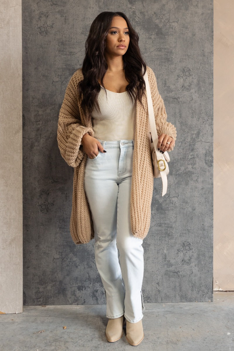 Full body front view of model wearing the Jayla Taupe Open Front Cardigan that has taupe cable knit fabric, an open front, dropped shoulders, a thigh length hem, and long bubble sleeves.