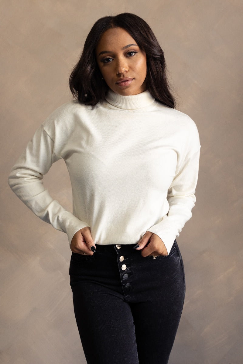 Front view of model wearing the Gabriela Cream Turtleneck Sweater which features cream knit fabric, ribbed trim, a turtleneck neckline, dropped shoulders, and long sleeves with cuffs.