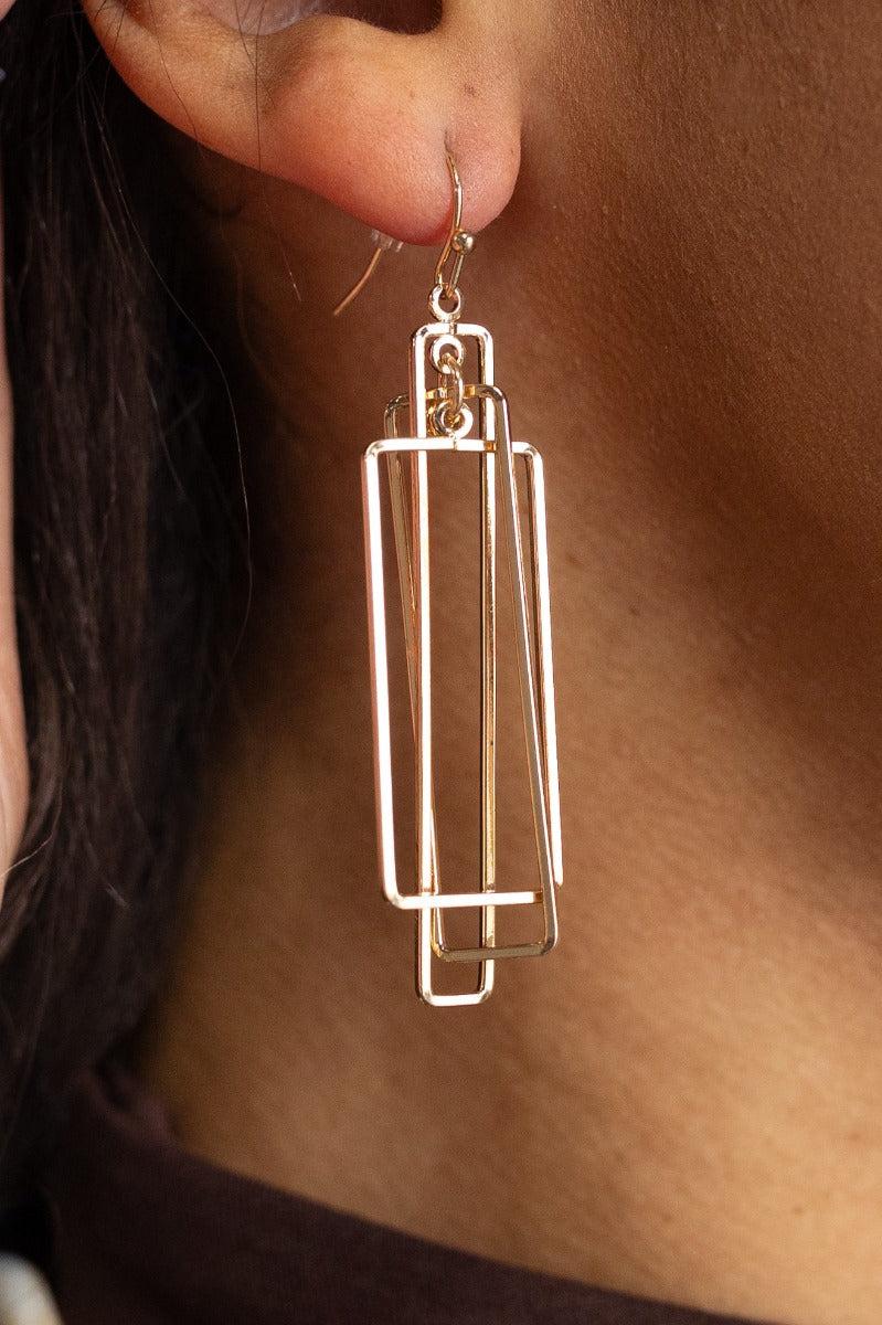 Close up side image of model wearing the Delilah Gold Geometric Earrings that feature layered gold rectangle pendants and a hook back.