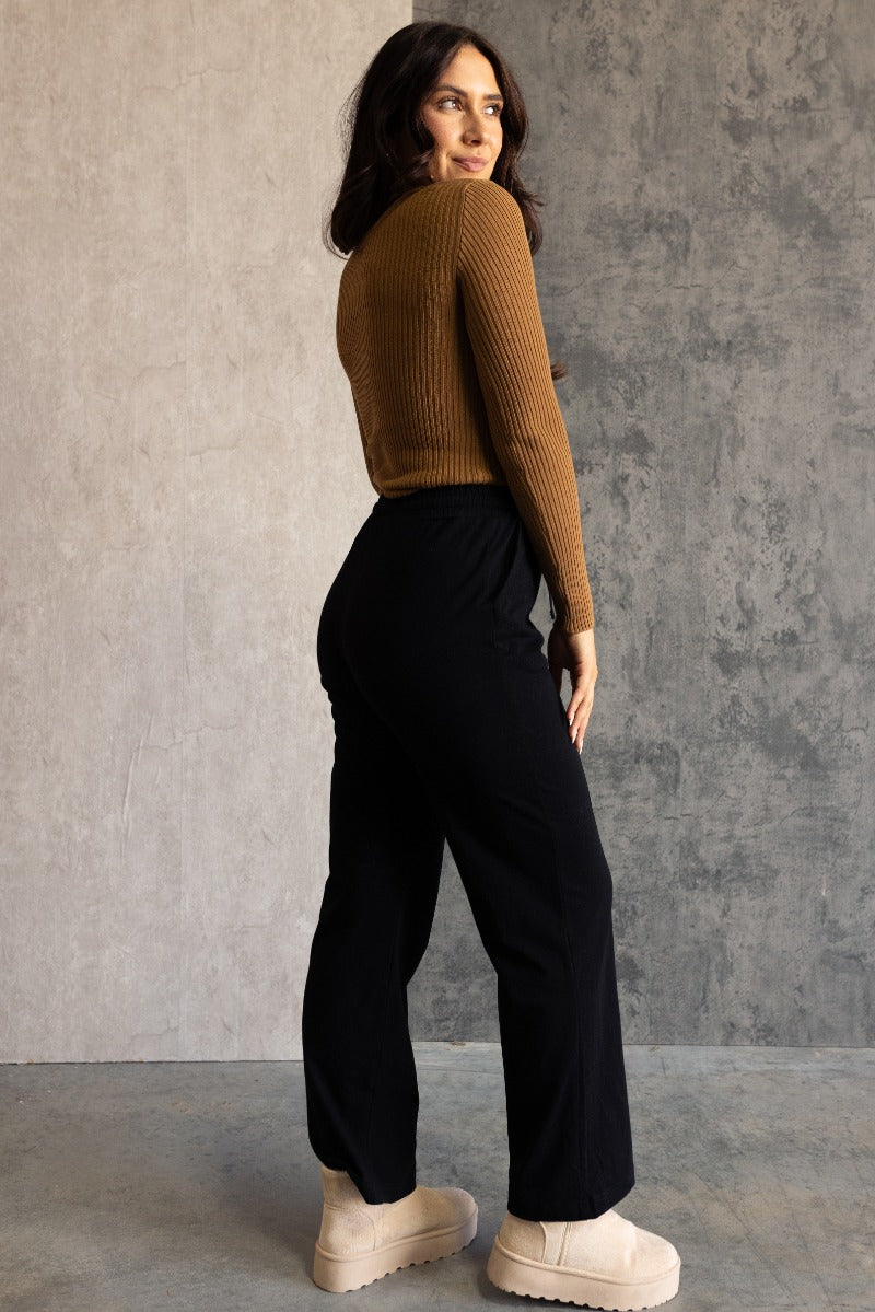 Full body back view of model wearing the Aubrey Black Lounge Pants which features black cotton fabric, two front pockets, an elastic waistband with drawstring ties, and wide legs.