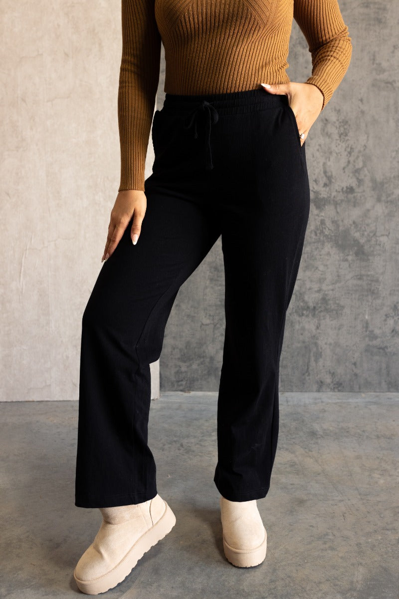 Front view of model wearing the Aubrey Black Lounge Pants which features black cotton fabric, two front pockets, an elastic waistband with drawstring ties, and wide legs.