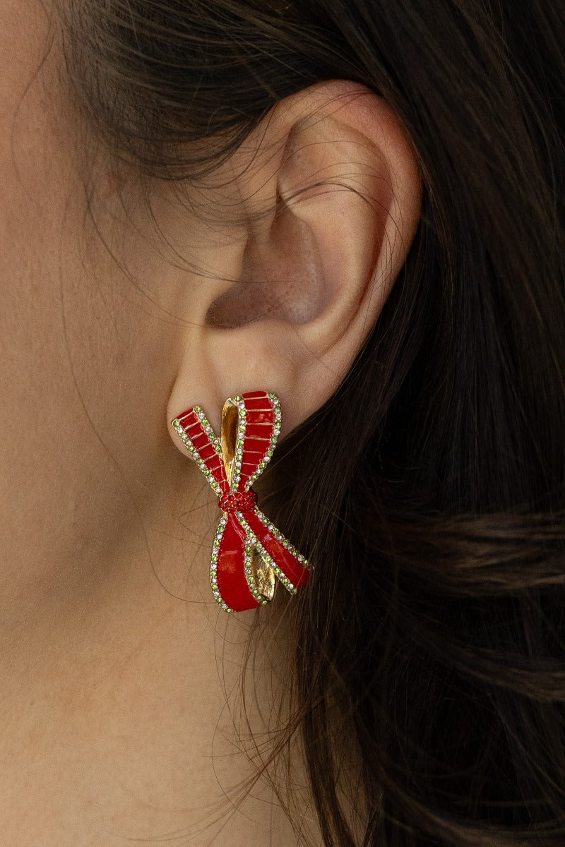 Close-up side view of model wearing the Noel Red Bow Earrings that feature red bow-shaped studs with red, light green and clear rhinestones and set in gold.