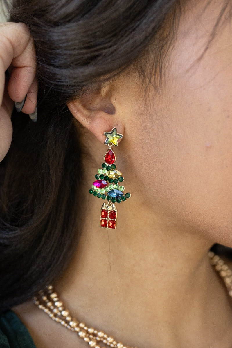 Side view of model wearing the Christmas Tree Multi-Color Stone Earrings that have multi-colored stones forming a Christmas Tree shape with green stars on post backs and red gift pendants.