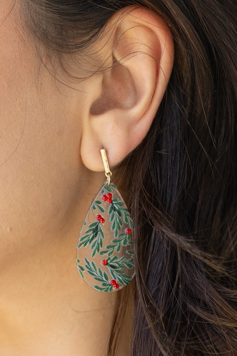 Close-up view of model wearing the Holly Teardrop Earrings that have clear teardop pendants with green holly designs, red jewel details, and brushed gold hardware.