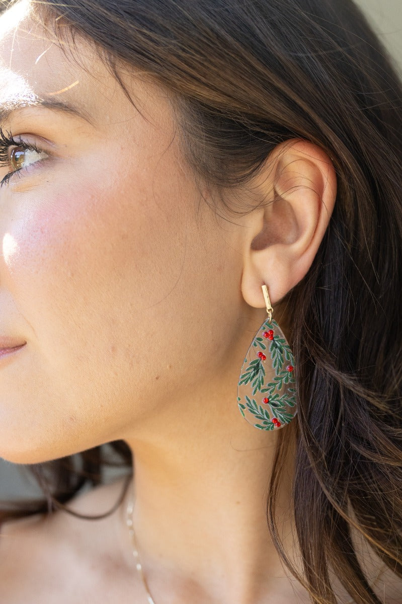 Side view of model wearing the Holly Teardrop Earrings that have clear teardop pendants with green holly designs, red jewel details, and brushed gold hardware.
