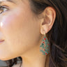 Side view of model wearing the Holly Teardrop Earrings that have clear teardop pendants with green holly designs, red jewel details, and brushed gold hardware.