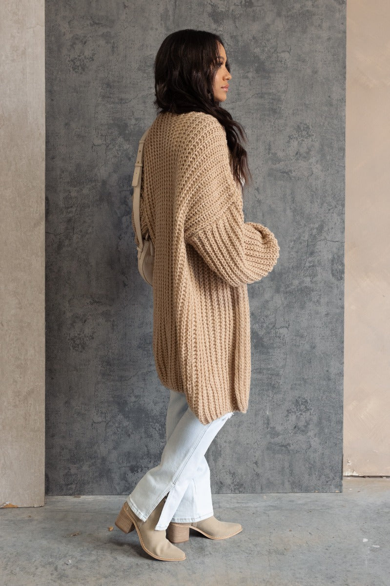 Full body side view of model wearing the Jayla Taupe Open Front Cardigan that has taupe cable knit fabric, an open front, dropped shoulders, a thigh length hem, and long bubble sleeves.