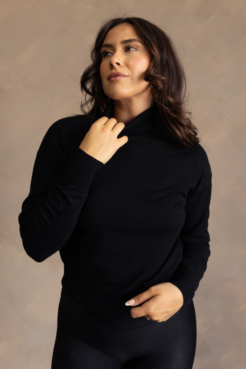 Front view of model wearing the Gabriela Black Turtleneck Sweater that has black knit fabric, ribbed trim, a turtleneck neckline, dropped shoulders, and long sleeves with cuffs.
