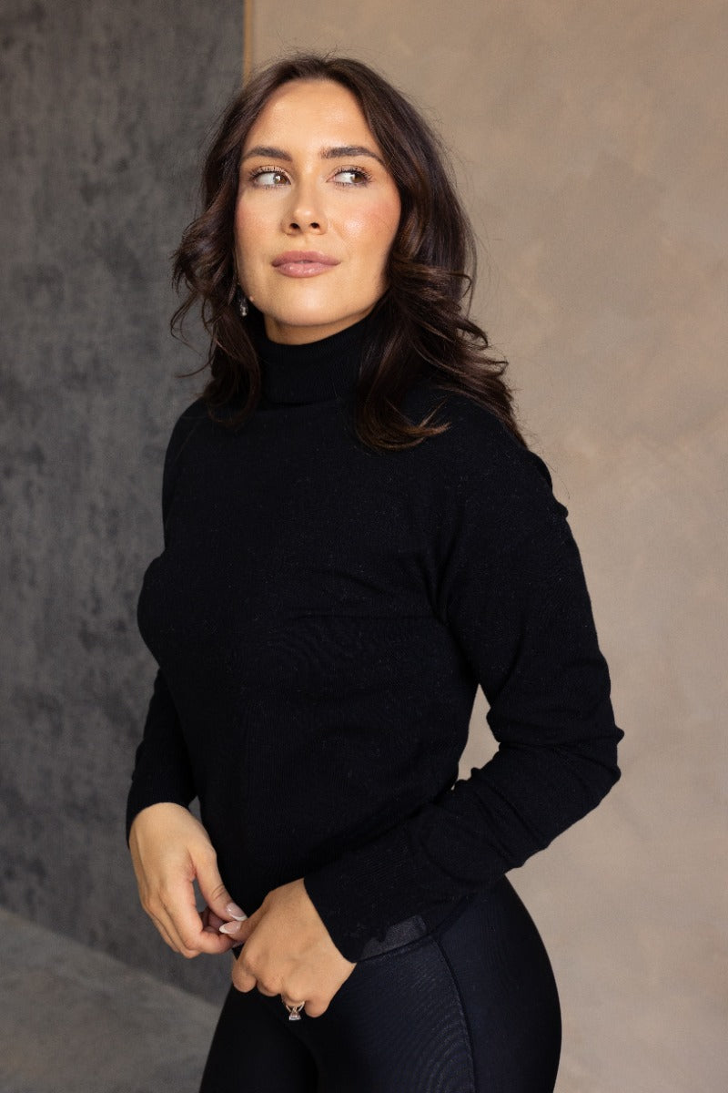 Side view of model wearing the Gabriela Black Turtleneck Sweater that has black knit fabric, ribbed trim, a turtleneck neckline, dropped shoulders, and long sleeves with cuffs.