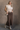 Full body view of model wearing the Angela Brown Front Slit Midi Skirt which features brown cotton fabric, midi length, front slit detail, front zipper with button closure, belt loops, two front pockets, two back pockets and fray hem.