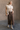 Full body view of model wearing the Angela Brown Front Slit Midi Skirt which features brown cotton fabric, midi length, front slit detail, front zipper with button closure, belt loops, two front pockets, two back pockets and fray hem.