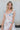 Front view of model wearing the In Full Bloom Dress that has ivory fabric with a pink, red, yellow and teal floral pattern, a scoop neckline with ruffle details, ruffle straps, and a smocked cut-out back.