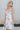 Frontal side view of model wearing the In Full Bloom Dress that has ivory fabric with a pink, red, yellow and teal floral pattern, a scoop neckline with ruffle details, ruffle straps, and a smocked cut-out back.