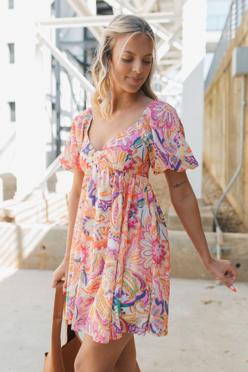 Frontal side view of model wearing the Under The Cabana Dress that has blue, green, orange, pink and yellow fabric with a floral pattern, ivory lining, a sweetheart neck, an elastic waist, a smocked back, and short bubble sleeves